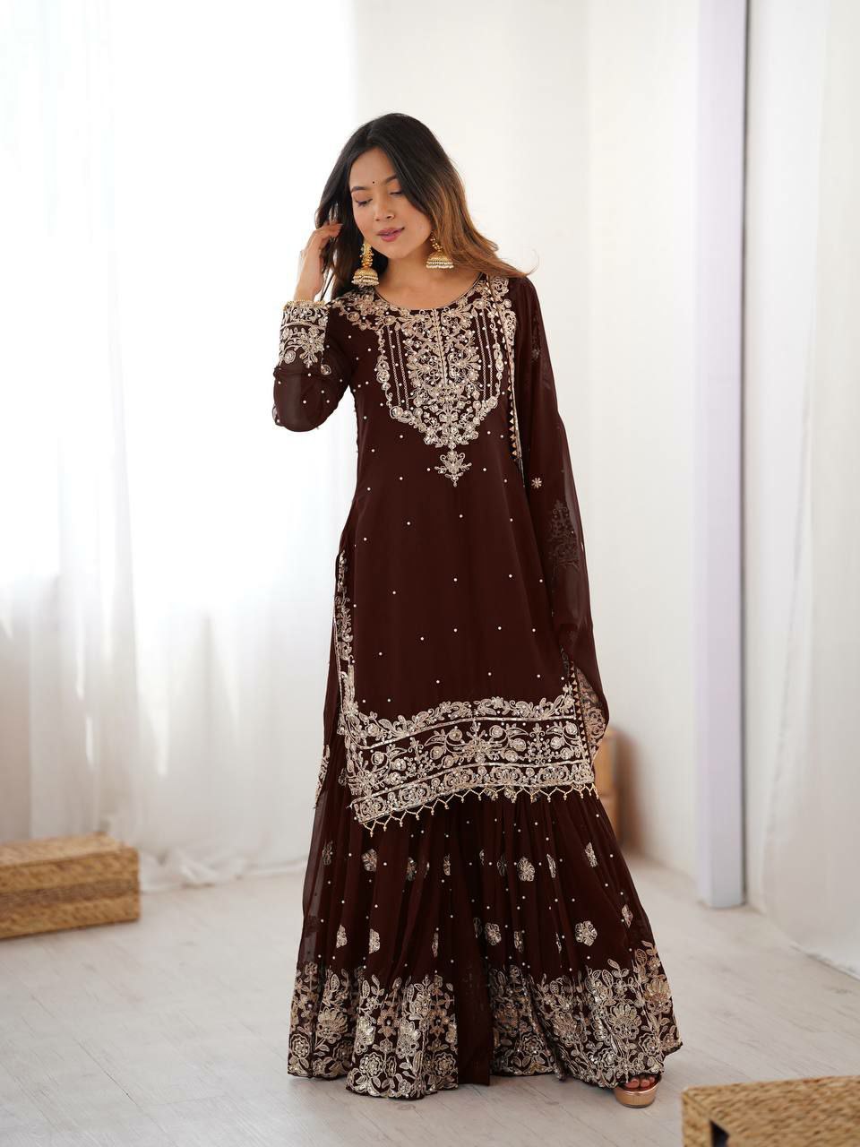 Woman wearing a long brown dress with white embroidery in a minimal indoor setting.